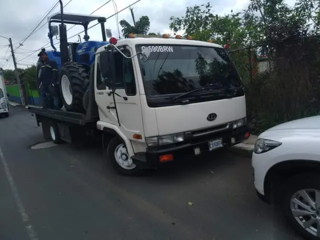 Camión de plataforma blanco transportando un tractor azul en la calle
